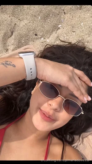 Woman with dark curly hair wearing red bikini top and sunglasses lying on sand.