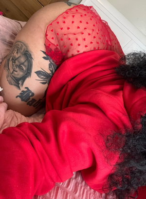 Woman with curly hair and tattoos on back, wearing red hoodie and sheer red heart-patterned fabric,