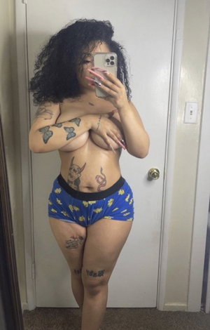 Woman with curly hair and tattoos wears blue patterned shorts, posing in front of a door.
