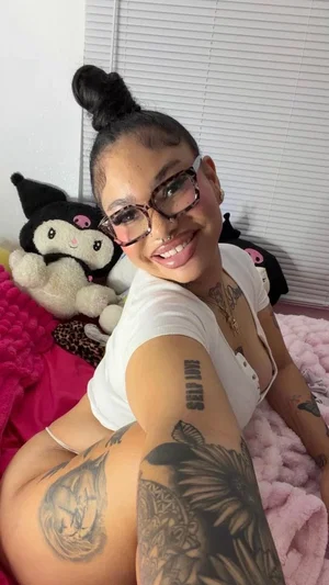 Woman with tattoos and glasses in white top, sitting on pink blanket with stuffed toys.