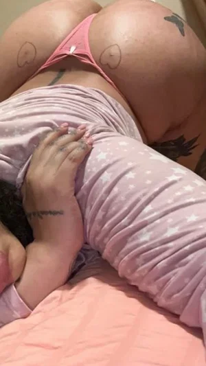 Woman in pink underwear and star-patterned top lying on bed, showing tattoos on hips and hand.