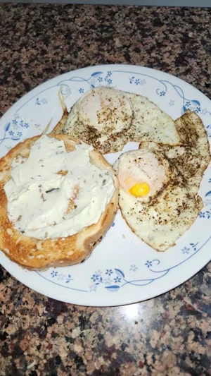 Plate with bagel and cream cheese, two fried eggs on granite countertop.