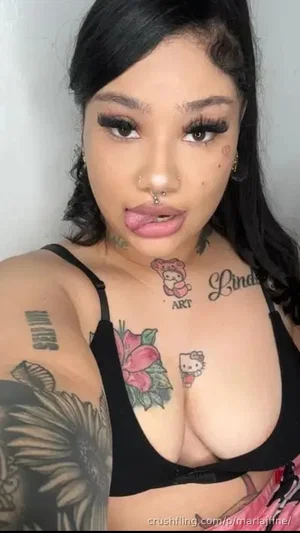Woman with tattoos, black bra, and dark hair posing indoors.