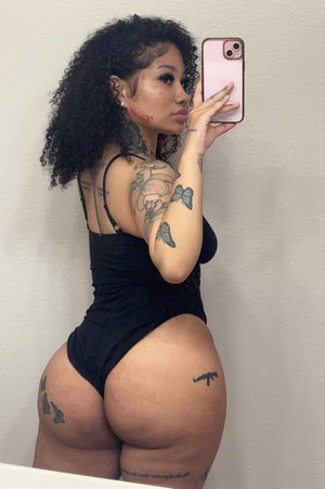 Woman with curly hair and tattoos wears black bodysuit, takes mirror selfie indoors.