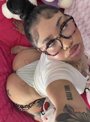Woman with tattoos and glasses lying on pink bedding, wearing striped top and leopard underwear.