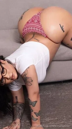Woman with tattoos in pink lace underwear and white crop top, posing on a couch.
