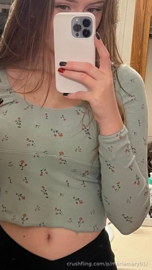 Woman in floral crop top and black bottoms taking mirror selfie indoors.