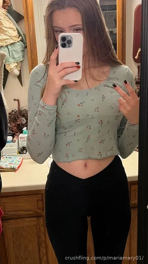 Woman in floral crop top and black pants taking mirror selfie in bathroom.