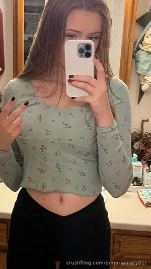 Woman in floral crop top and black pants taking mirror selfie in bathroom.