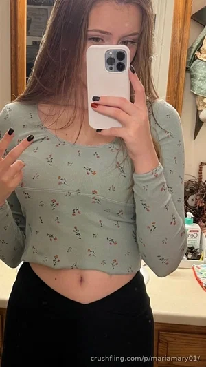 Woman in floral crop top and black pants taking mirror selfie in bathroom.