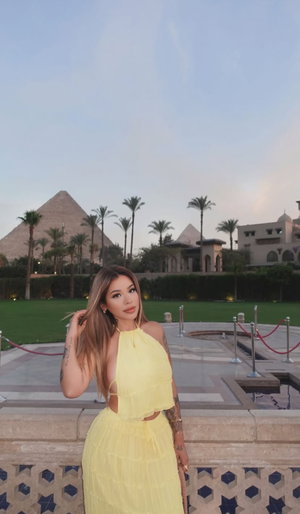 Blonde woman in yellow halter top and skirt outdoors near pyramids.