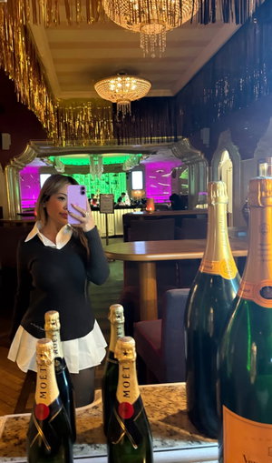 Woman in black sweater and white skirt taking selfie in bar.
