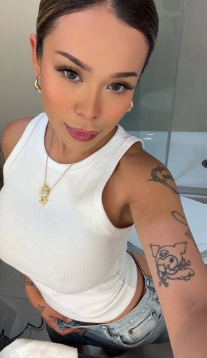 Woman with tattoos in white tank top taking bathroom selfie.