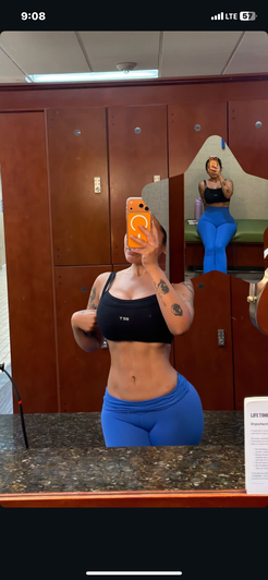 Woman in black top and blue leggings taking gym mirror selfie.