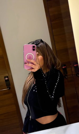 Woman with long hair in black crop top and pants, taking mirror selfie indoors.