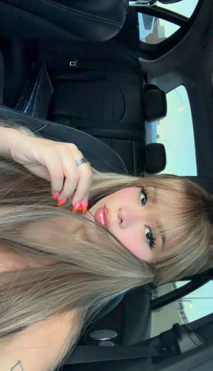 Woman with long blonde hair and bright nails posing in a car, showing shoulder and arm.