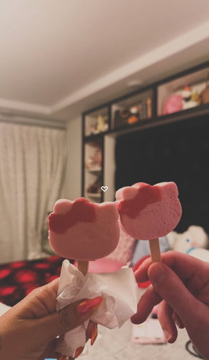 Hands holding pink Hello Kitty-shaped popsicles in a bedroom setting.