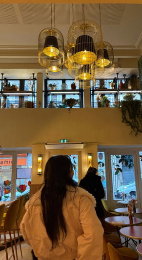 Woman with long dark hair in a cream coat standing in a cafe with hanging lights and plants.