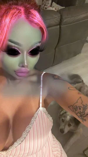 Person with pink hair and green face makeup wears striped camisole, indoors with dog nearby.