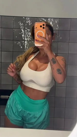 Woman in white crop top and teal shorts takes mirror selfie in tiled room.