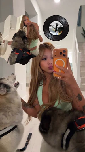 Woman with long hair and tattoos in light green top takes mirror selfie with dogs indoors.