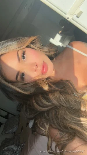 Woman with wavy hair in a white top, taking a selfie indoors near a bed.
