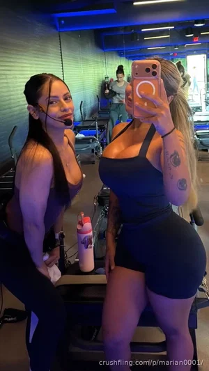 Two women in athletic wear pose for a mirror selfie in a gym.