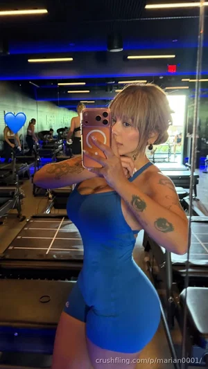 Woman in blue workout outfit with tattoos takes mirror selfie in gym.