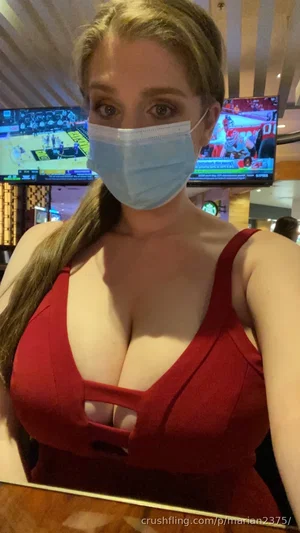 Woman in red top with cutout, wearing face mask, indoors with TVs in background.