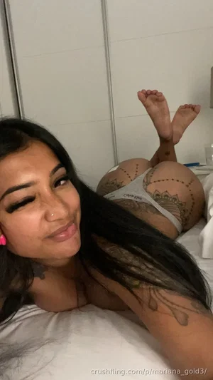 Woman with long dark hair and tattoos lying on bed, wearing white underwear, indoor setting.