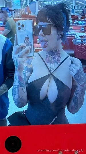 Woman with tattoos and sunglasses in a store, wearing a revealing black top, taking a mirror selfie.