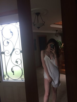 Woman in white lace dress taking mirror selfie indoors near decorative glass door.