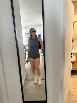 Woman in gray bodysuit and white sneakers takes mirror selfie in indoor setting.