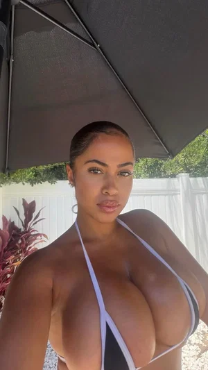 Woman in white bikini top under umbrella, outdoor setting, curvy figure.