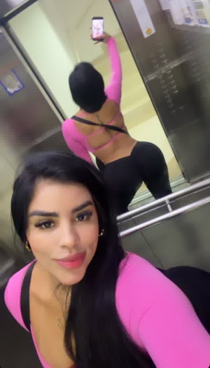Woman with long black hair in pink top and black pants, taking mirror selfie in elevator.