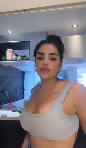 Woman in gray crop top posing in kitchen, dark hair in bun, visible tattoo on collarbone.
