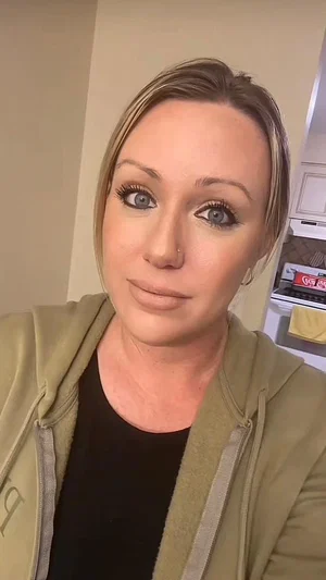 Woman with blonde hair and makeup wearing a green hoodie and black top indoors.