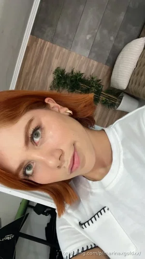 Woman with red hair in white t-shirt, indoor setting with plant and furniture.