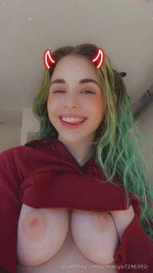 Woman with green hair, bare breasts covered by red hoodie, smiling in indoor setting.