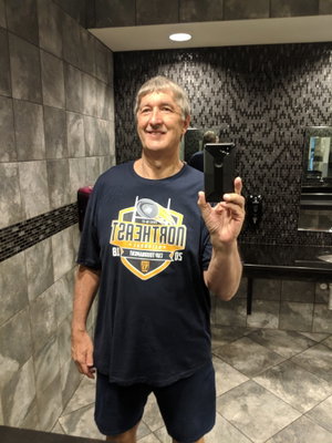 Man in dark t-shirt and shorts takes mirror selfie in tiled bathroom.