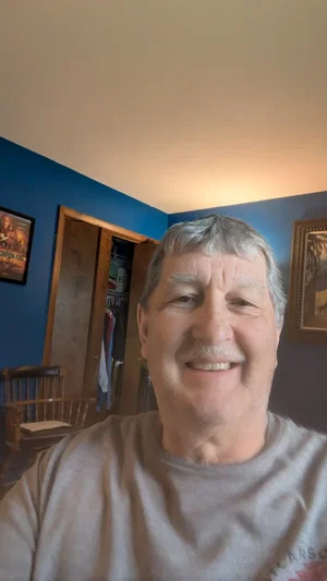 Smiling older man in gray t-shirt, indoors, blue walls, wooden chair visible.