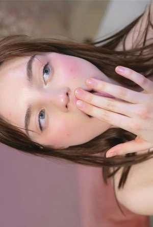 Woman with brown hair and blue eyes covering mouth with hand, close-up selfie in soft lighting.