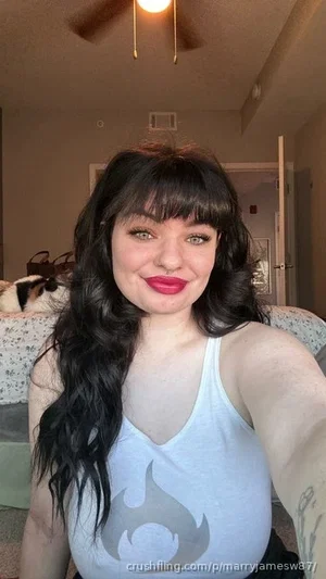 Woman with long dark hair and bangs in a white tank top, taking a selfie indoors.