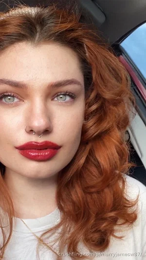 Woman with red curly hair and red lipstick, wearing white top, inside a car.