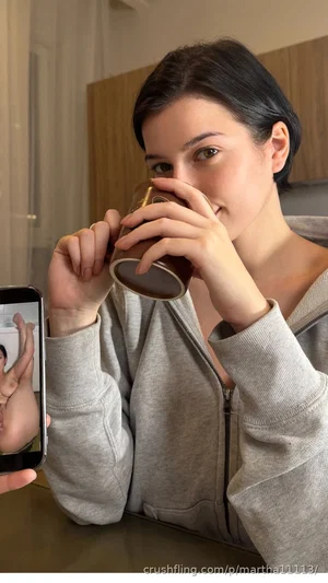 Woman with short dark hair in gray hoodie drinking from mug, holding phone for mirror selfie.