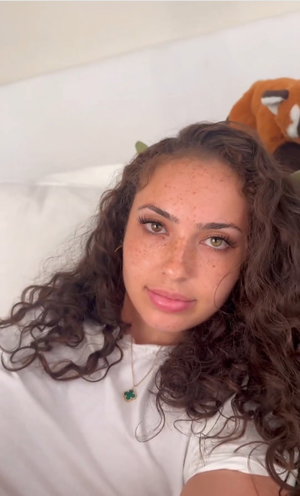 Woman with curly brown hair and freckles wearing white top, lying on bed with stuffed animal behind.