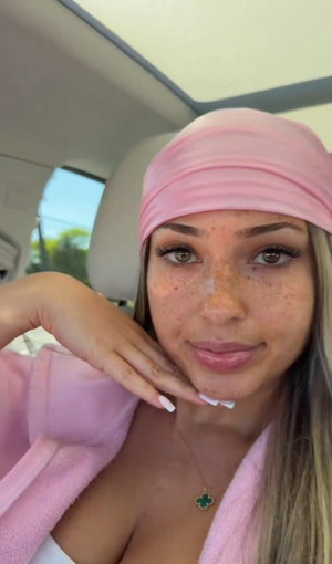 Woman with freckles and blonde hair in pink headband and robe, sitting in car.