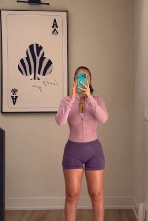 Woman in pink zip-up top and purple shorts takes mirror selfie in bedroom.