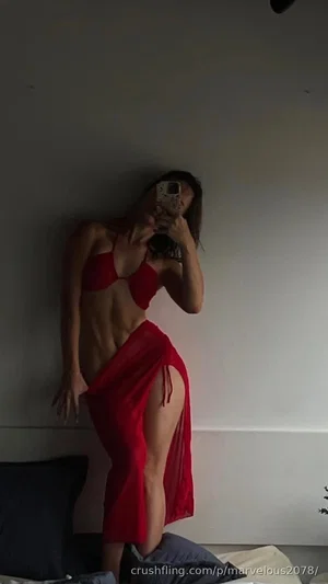 Woman in red lingerie and skirt poses against wall in bedroom, holding phone for mirror selfie.
