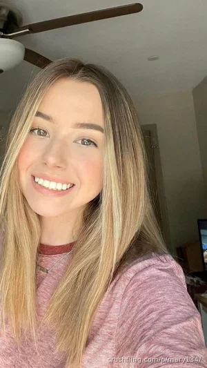 Woman with long blonde hair wearing a pink top, smiling in a bedroom selfie.